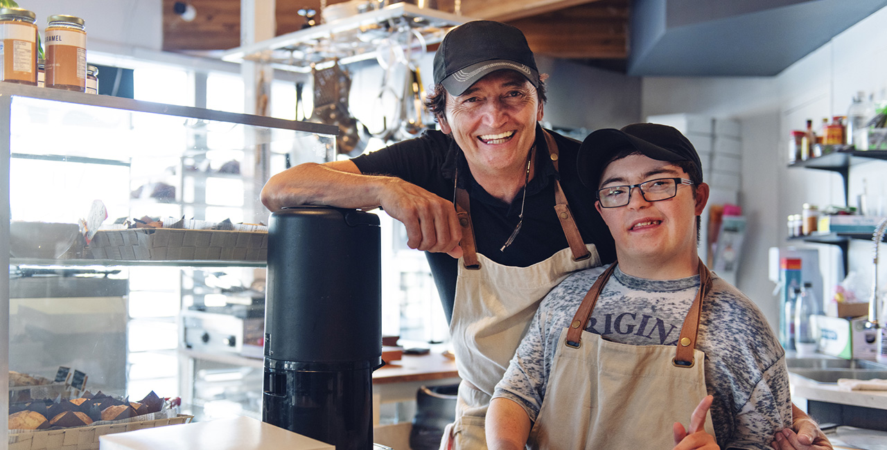 Junger freundlicher Mann mit Down-Syndrom und älterer Herr arbeiten gemeinsam in einem Café